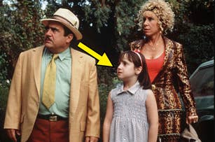 Danny DeVito, Mara Wilson, and Rhea Perlman dressed in vintage style, posing outdoors. DeVito in a hat, Wilson in a dress, Perlman in a leopard print outfit